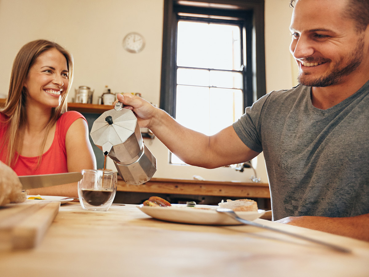 Man and woman sitting at a table, smiling, while the man pours a cup of coffee.